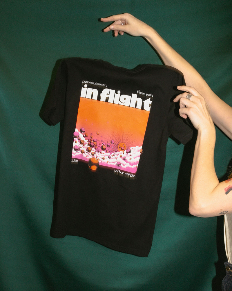 A person holds up a black short-sleeve graphic T-shirt against a dark green backdrop. The shirt features a large front print with the words "in flight" in white over a bright orange sky and pink clouds.